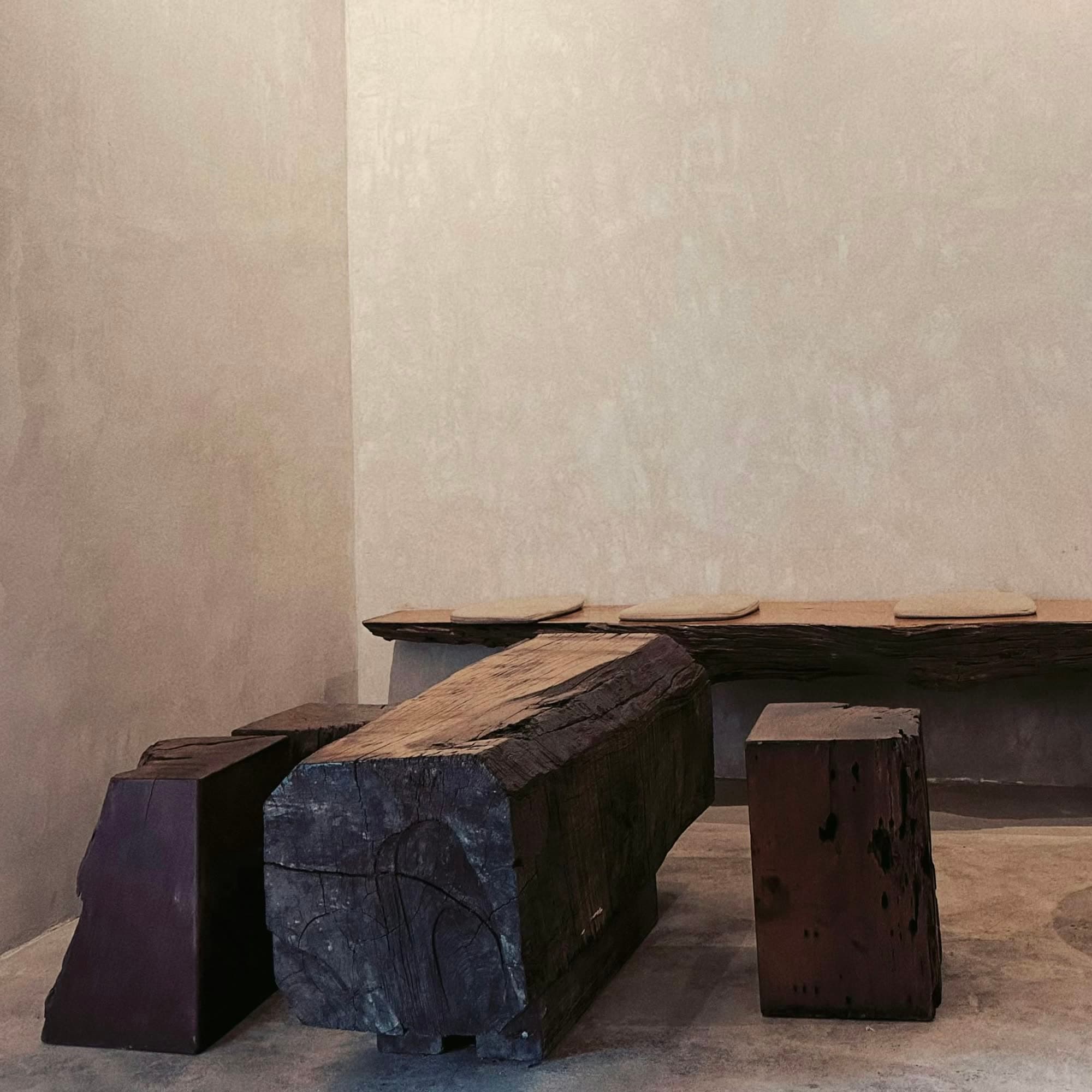 A plastered room in soft light, with a low bench and blocks of dark, weathered timber against the wall.