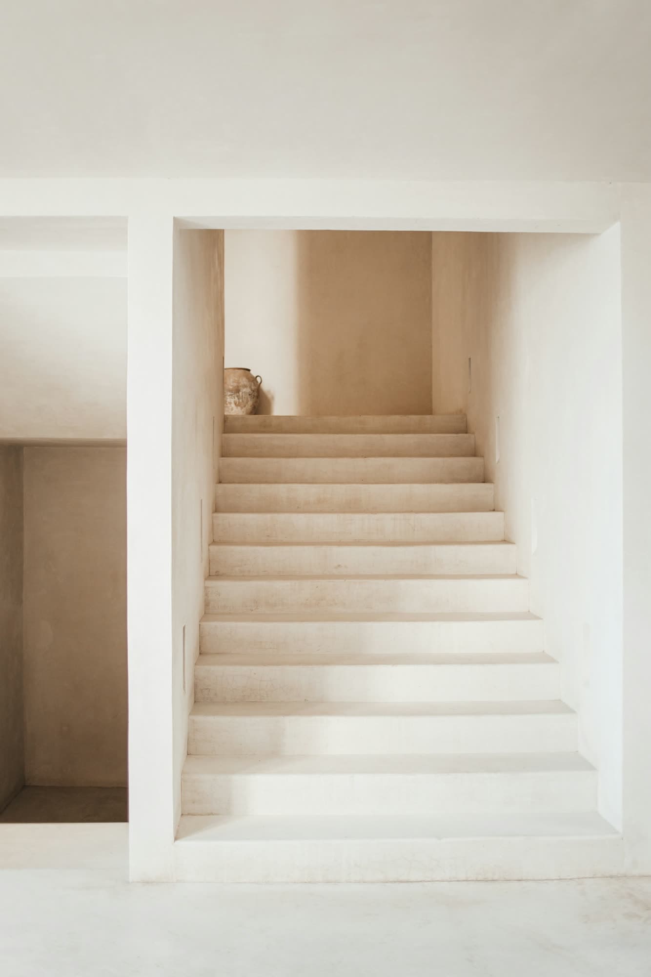 Smooth plaster steps rising between warm white walls towards a clay pot on the landing.