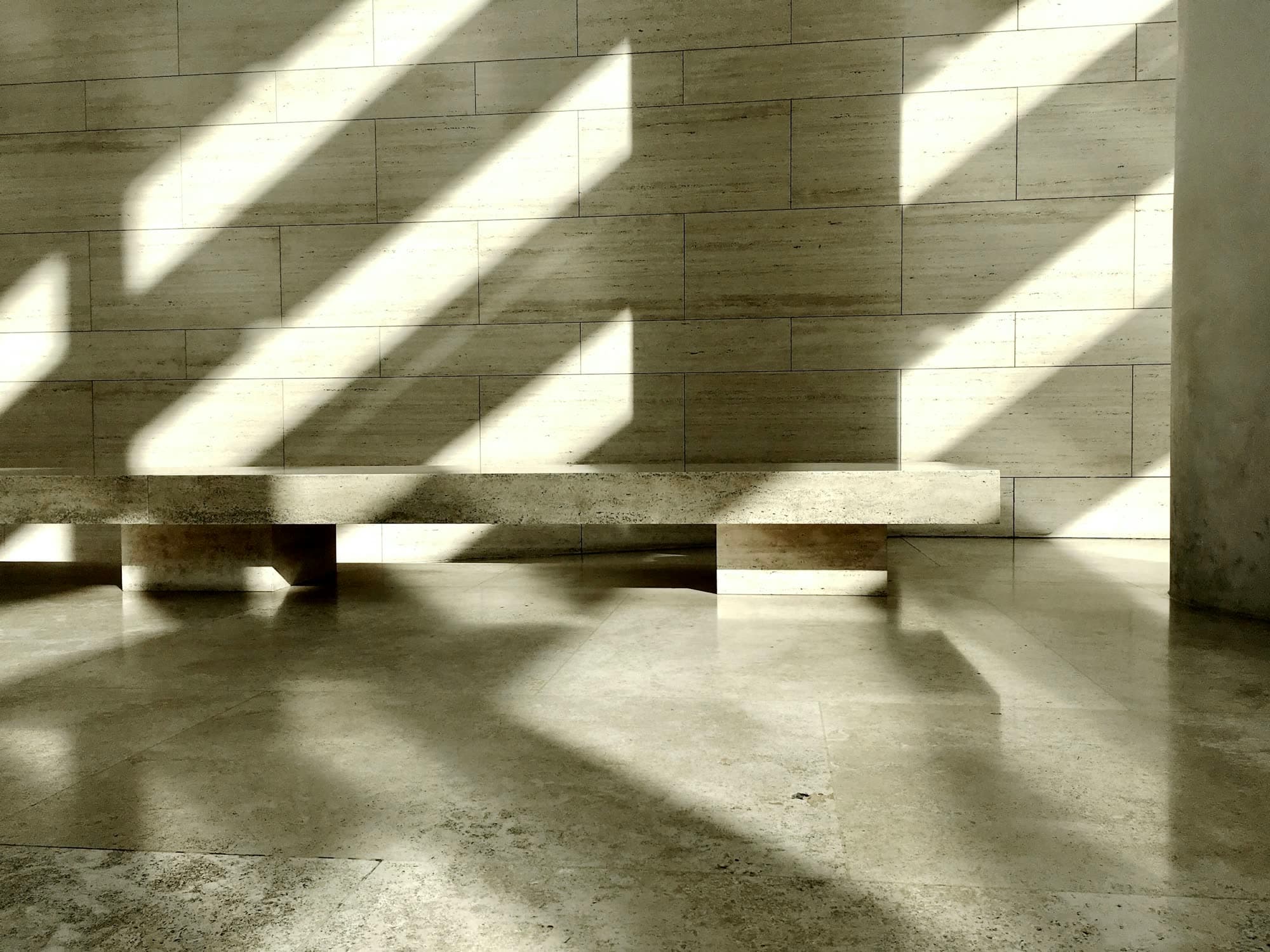 Bands of sunlight across a travertine wall and a long, low travertine bench.