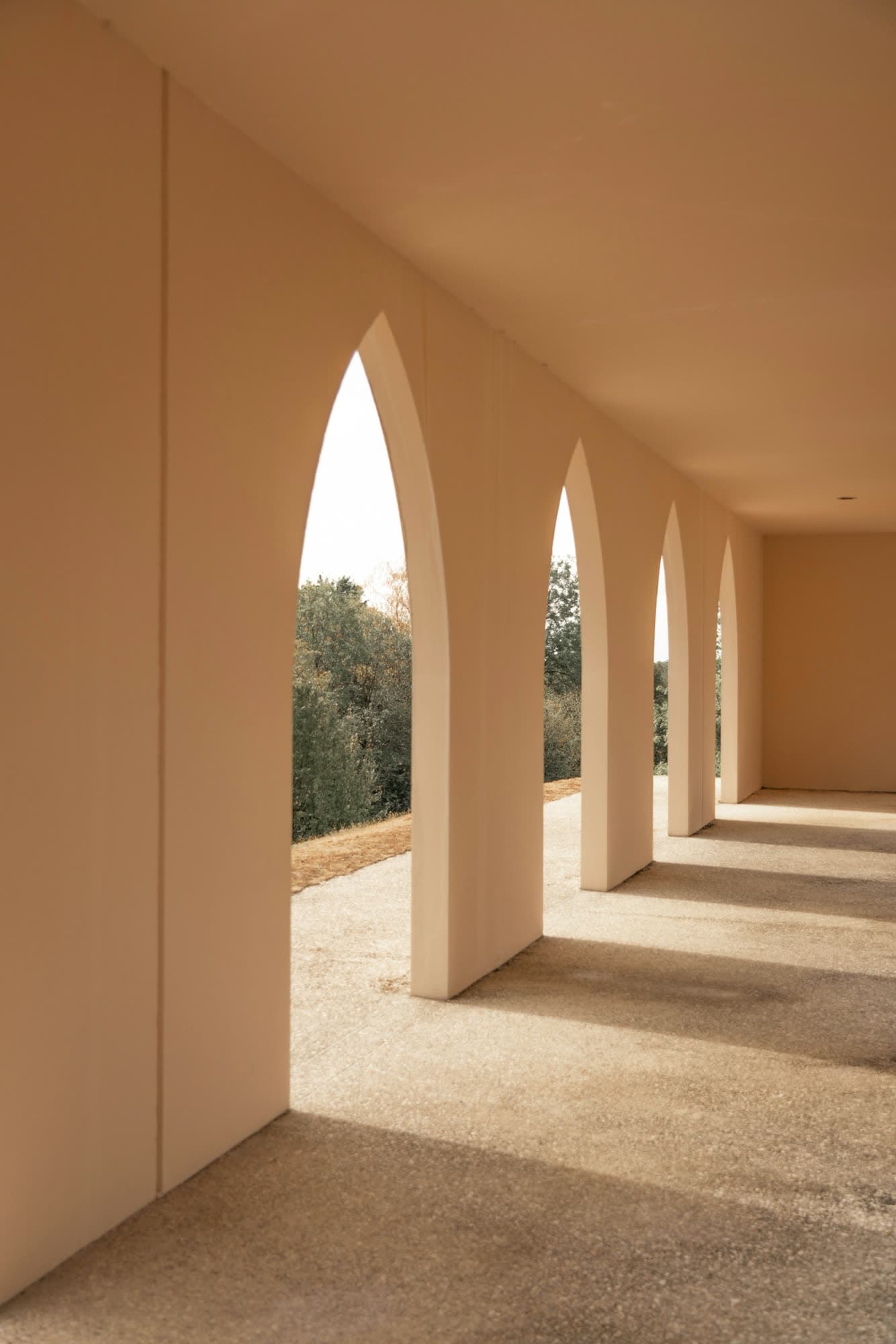 A sunlit arcade of tall pointed arches in warm plaster, looking out to trees.